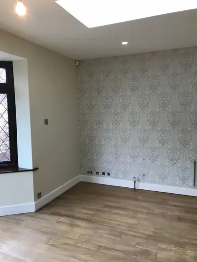 Empty room with a silver-grey damask wallpapered feature wall, pale green-grey painted walls, white skirting and a wood-effect floor.