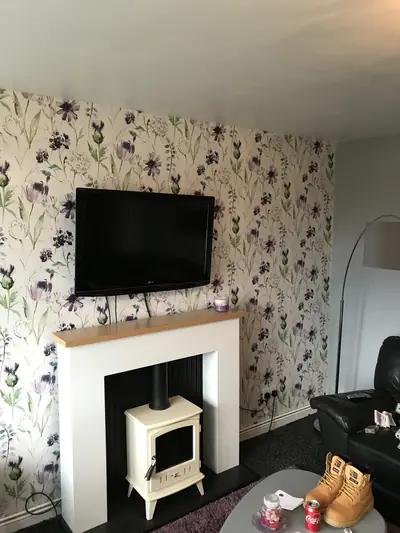 Living room chimney breast wall hung with cream floral wallpaper, a wall-mounted television above a white fire surround and cream stove.