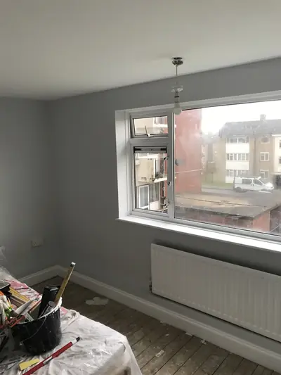 Bedroom with grey painted walls, white ceiling and skirting, a large window and radiator, with a paint bucket, dust sheet and bare floorboards in the foreground.