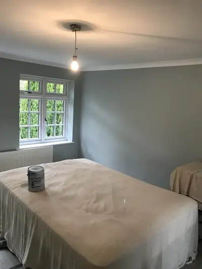 Bedroom painted in pale blue-grey emulsion with the bed covered by a cotton dust sheet and a paint tin standing on it.