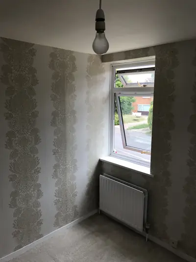 Small bedroom corner papered in a cream and gold damask stripe, with a white uPVC window, radiator and bare pendant light.