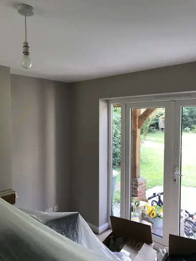 Room painted in warm grey with white French doors onto a garden, plastic sheeting over furniture and cardboard boxes on the floor.