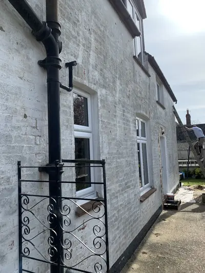 Cottage elevation part way through repainting, with patchy bare brickwork and a decorator working from a ladder at the far end.