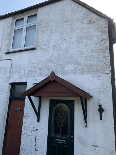 End elevation of a cottage in flaking, patchy white masonry paint, with a dark red timber door canopy and a weathered front door.