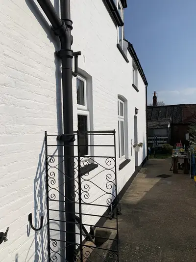 Side elevation of a house with white painted brickwork, black cast iron downpipe and dark window cills, photographed in sunlight.