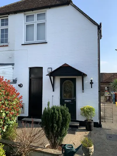 End of a cottage painted white with black doors, porch canopy and downpipe, in bright sunshine with shrubs in front.
