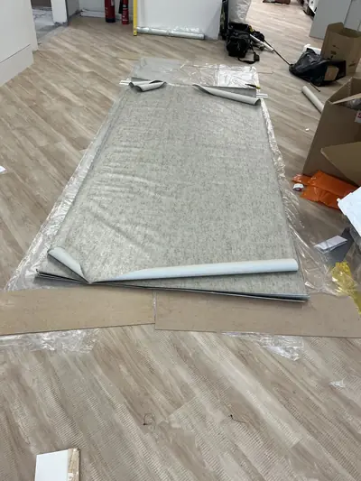 Lengths of grey textured wallcovering laid out on protective sheeting on a vinyl floor, with tool bags and packaging around them.