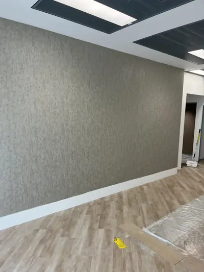 Office wall hung with textured greige vinyl wallcovering above a white skirting, with wood-effect vinyl flooring and a dark ceiling raft above.