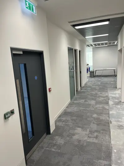 Office corridor with white painted walls and ceiling, a dark grey fire door with vision panel, green exit sign and grey carpet tiles.