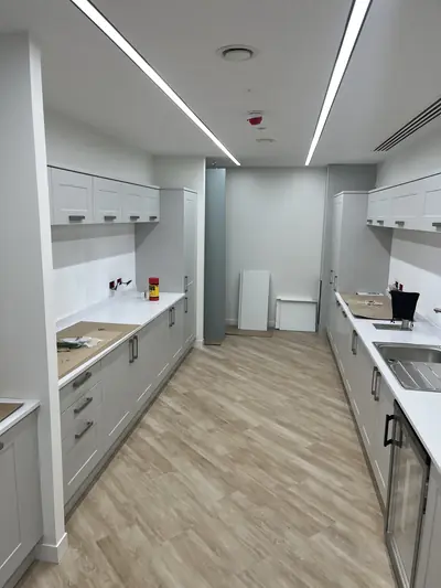 Office tea point with white painted walls and ceiling, grey shaker units both sides and wood-effect flooring, with protective card on the worktops.