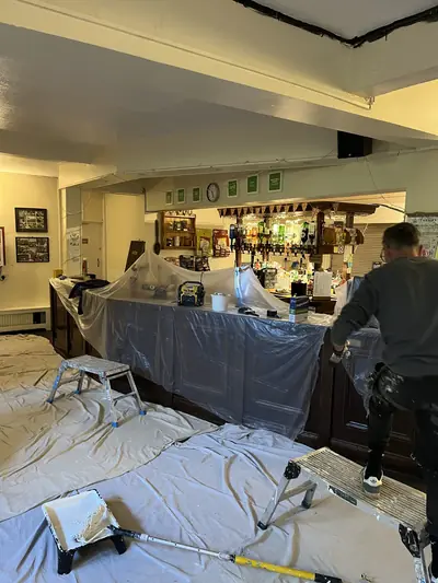 Pub bar area sheeted in polythene and dust sheets while the ceiling and walls are painted, with a decorator on a hop-up and a roller tray on the floor.