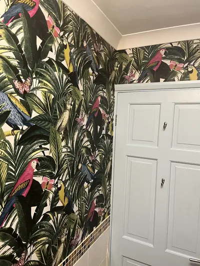 Corner of a bathroom papered with tropical parrot and foliage wallpaper, meeting a pale blue painted panel door and a white ceiling.