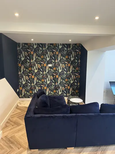 Living room with a dark botanical and bird wallpaper feature wall framed by navy painted walls, a navy sofa in the foreground and herringbone flooring.