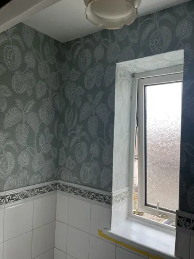Bathroom corner papered in green leaf-pattern wallpaper above a white tiled dado, with masking tape still on the painted window cill.